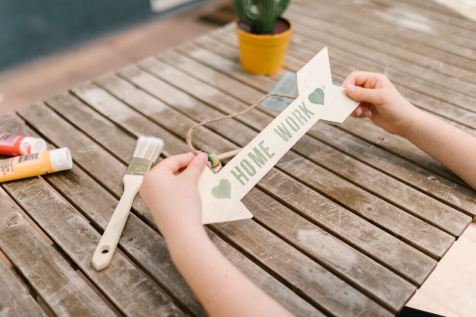 Hands creating a homework sign on a rustic wooden table with paints and brushes. (Photo by RDNE Stock project on Pexels)