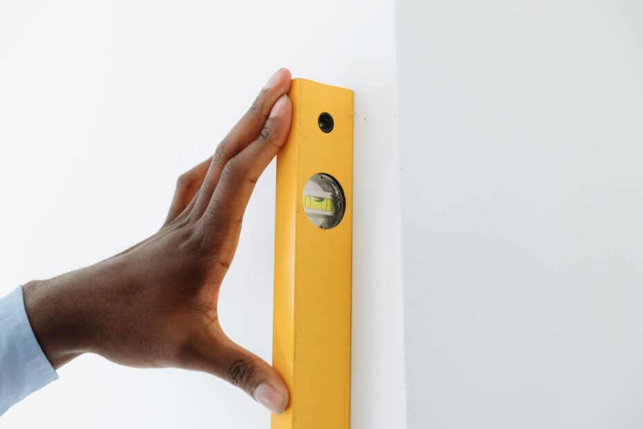 A close-up of a hand using a yellow level tool against a white wall to ensure vertical alignment. (Photo by Thirdman on Pexels)