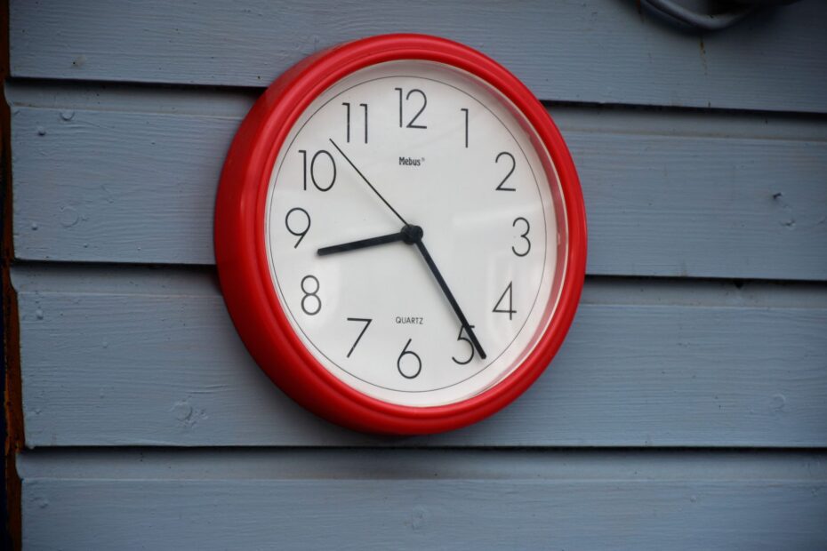 A red wall clock with black hands on a blue wooden wall, captured in daylight. (Photo by Dariusz Sbirenda on Pexels)