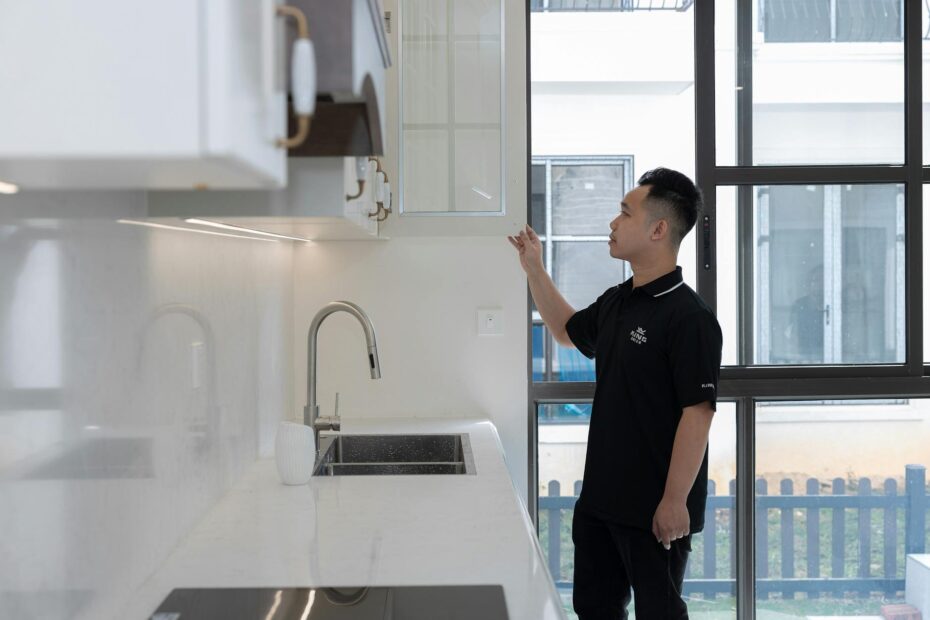 A man in black examines a kitchen cabinet inside a modern apartment with large windows. (Photo by HONG SON on Pexels)
