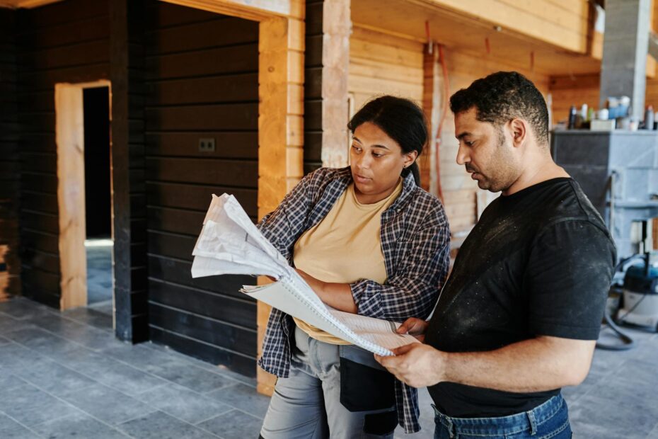 Two construction workers review blueprints inside a wooden structure, discussing plans. (Photo by Mikael Blomkvist on Pexels)