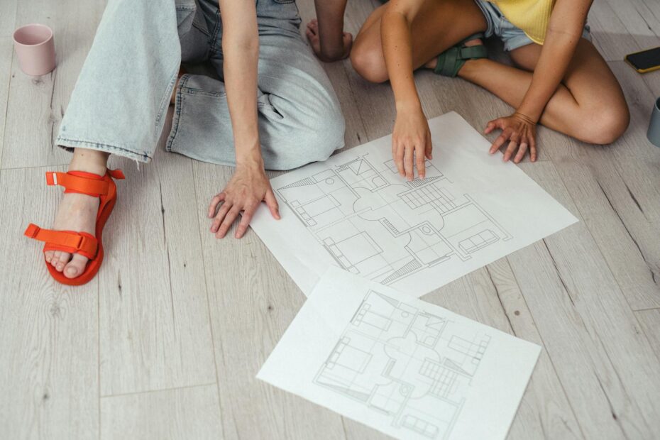 Two people discussing house blueprints on a light wooden floor. (Photo by SHVETS production on Pexels)